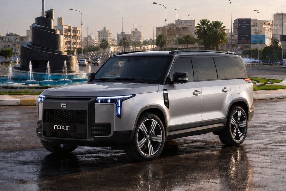 A silver Rox 01 SUV parked on a wet road beside a fountain, seen from the front left with its LED headlights on