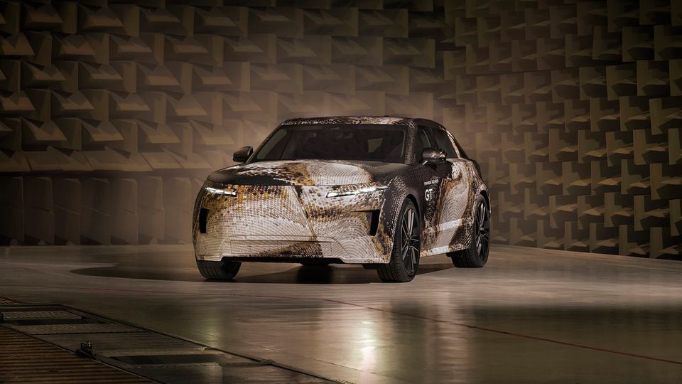 A Range Rover GT prototype SUV covered in camouflage wrap, seen from the front three-quarter angle inside what appears to be an acoustic testing facility