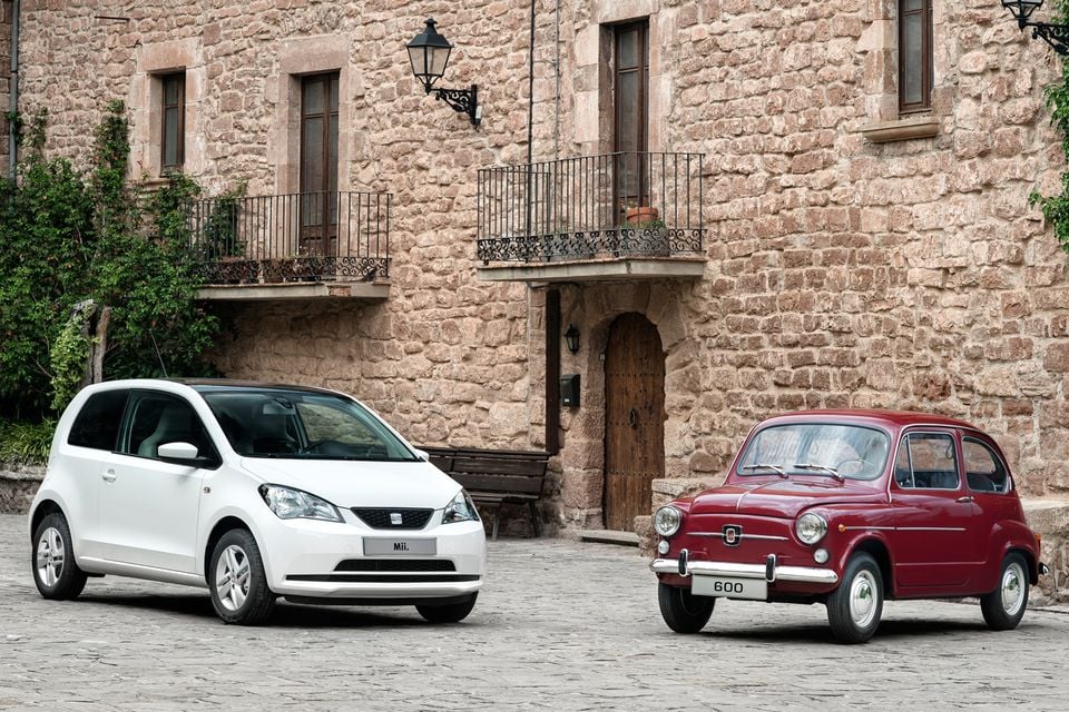 A white SEAT Mii hatchback parked alongside a classic red SEAT 600 on a cobblestone street in front of a stone building