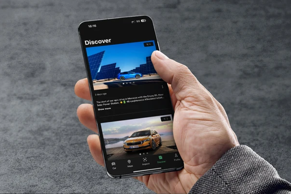 A hand holding a smartphone displaying a Skoda connected services app with car discovery content and vehicle images on screen