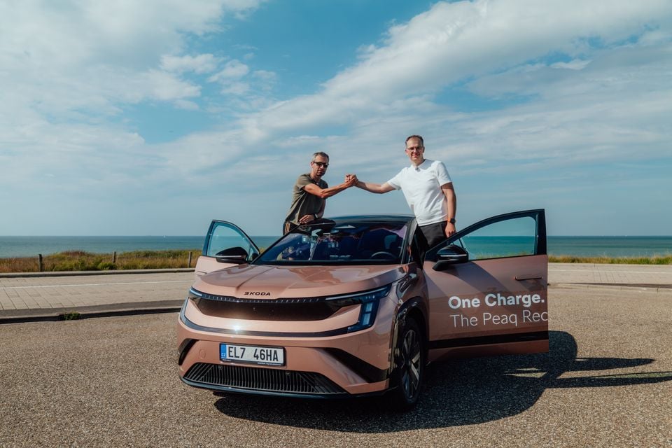 Two men shake hands over the bonnet of a bronze Skoda Peaq electric SUV parked near a coastline, with 'One Charge. The Peaq Rec...' branding visible on the door