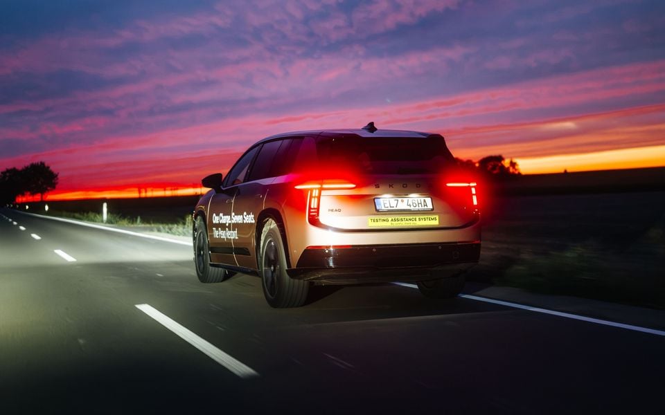 A Skoda Peaq electric SUV seen from the rear while driving on a road at sunset, with branding reading 'One Charge. Seven Seats. The Peaq Record.'
