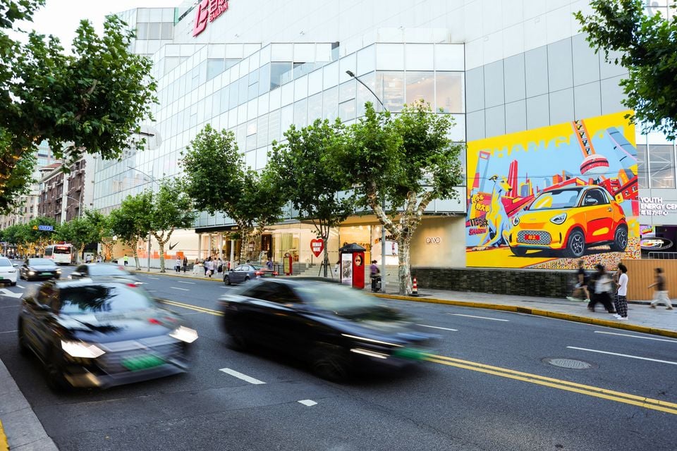 A colourful street mural on a building in Shanghai featuring a yellow Smart electric city car, with traffic passing in the foreground.