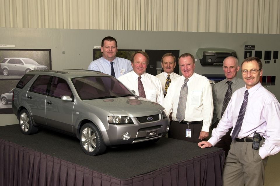 A silver Ford Territory SUV design model displayed on a stand with a group of six men standing beside it.