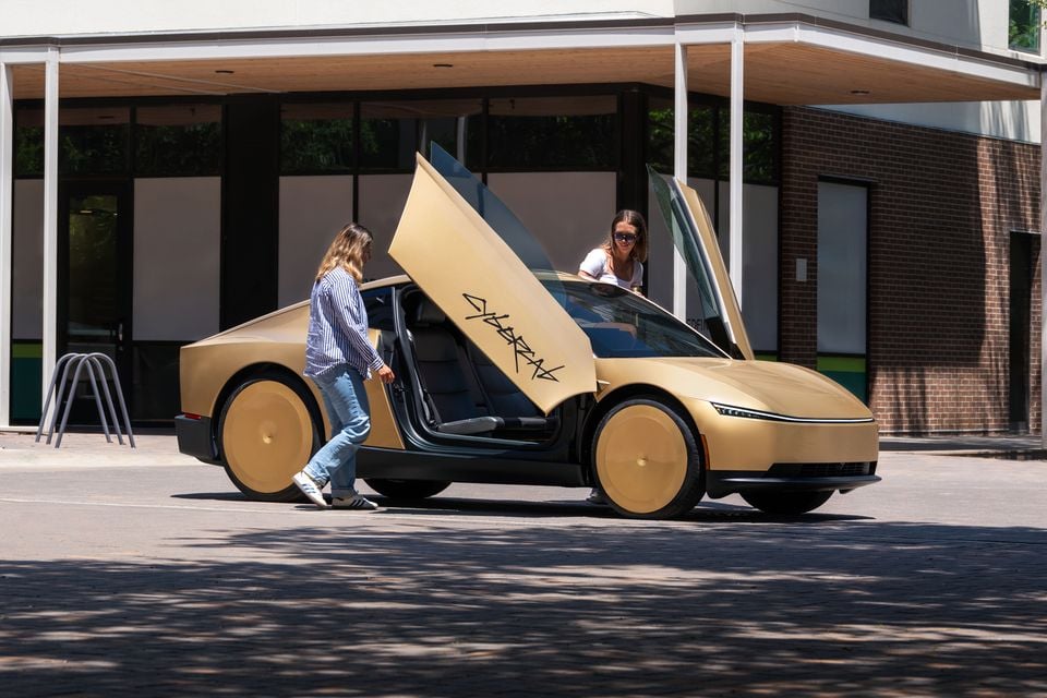 A gold Tesla Cybercab concept car with butterfly doors open, parked on a street, with two people approaching and entering the vehicle.