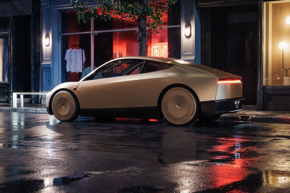 A Tesla Cybercab concept car in a champagne/gold finish parked on a wet city street at night, seen from the rear side angle