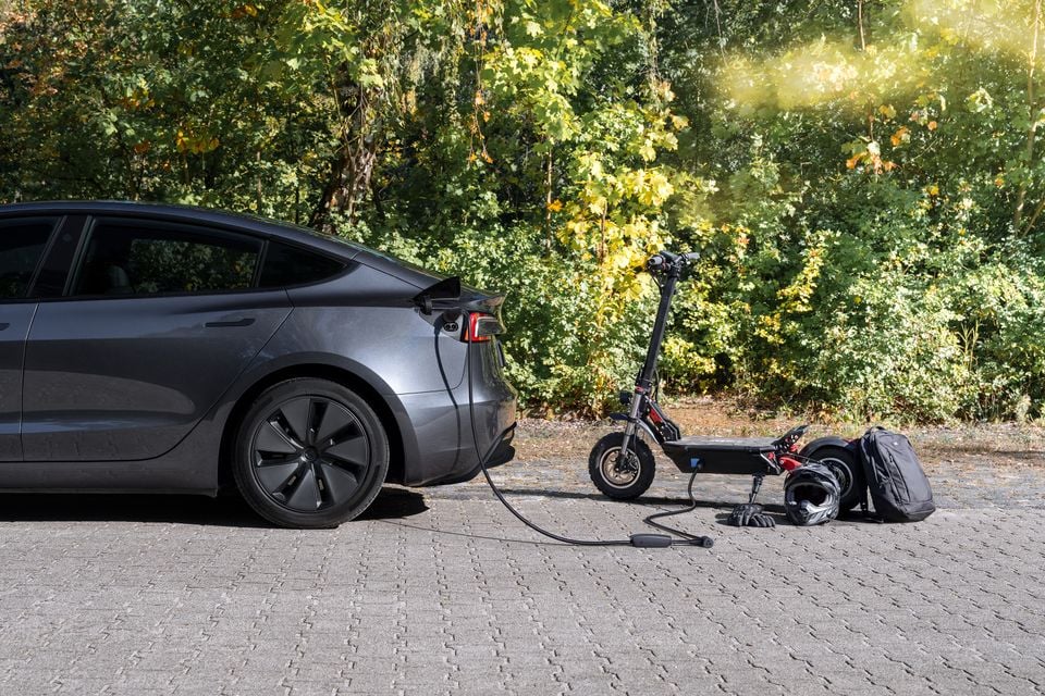 A grey Tesla Model 3 parked rear-facing on a paved area, using its Powershare feature to charge an electric scooter via a cable.