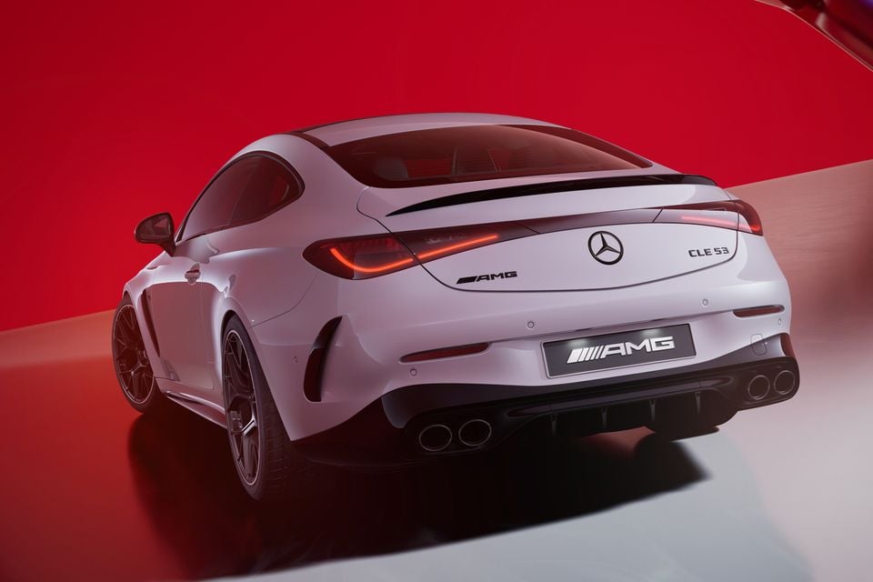 Rear three-quarter view of a white Mercedes-AMG CLE 53 coupe against a red studio background, showing AMG badging, quad exhausts, and LED tail lights