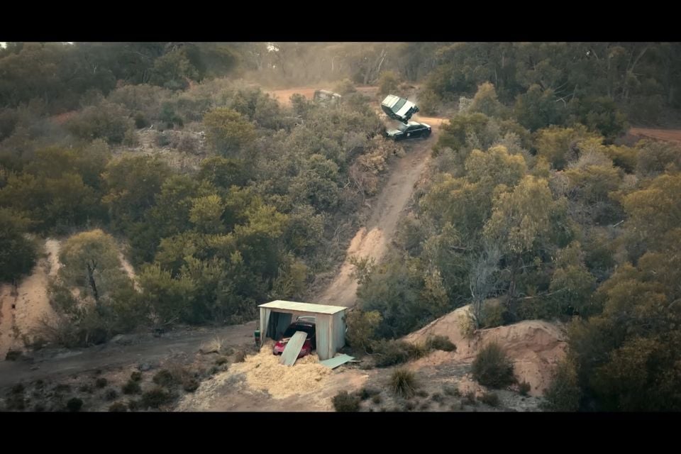 Aerial drone shot of vehicles on a dusty dirt track through Australian scrubland, with a small corrugated iron shed visible in the foreground