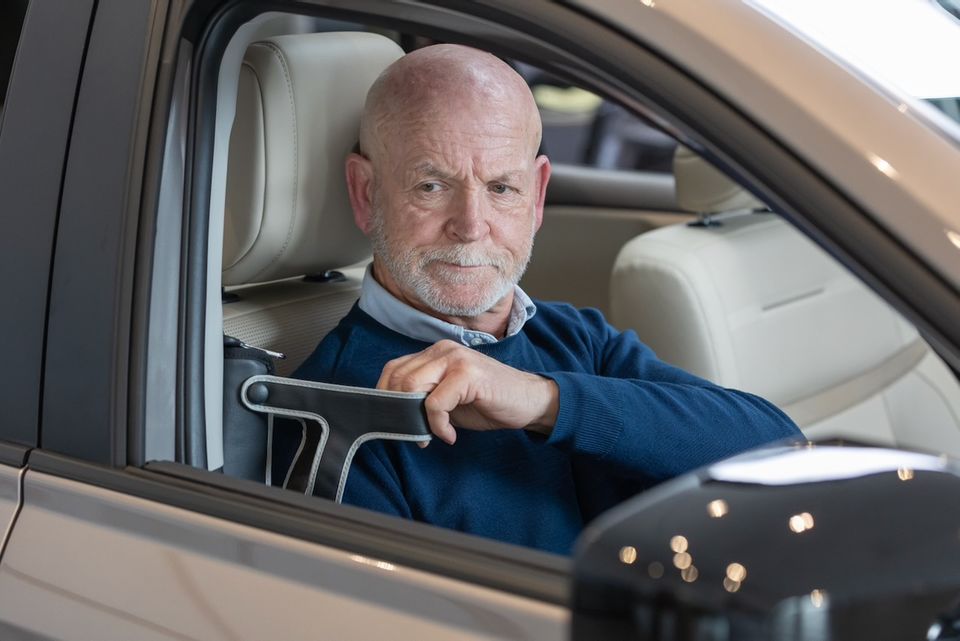 An older man sitting in the driver's seat of a car, gripping a mobility assist handle attached to the door frame, photographed from outside the vehicle.