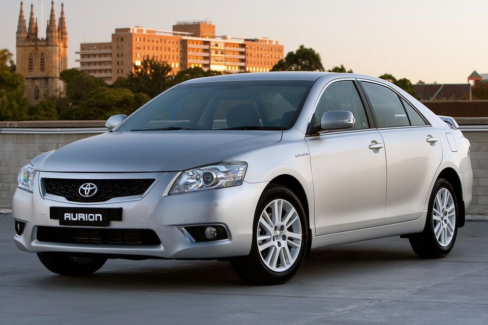 A silver Toyota Aurion sedan seen from the front-side angle, parked on a rooftop with a city backdrop at dusk