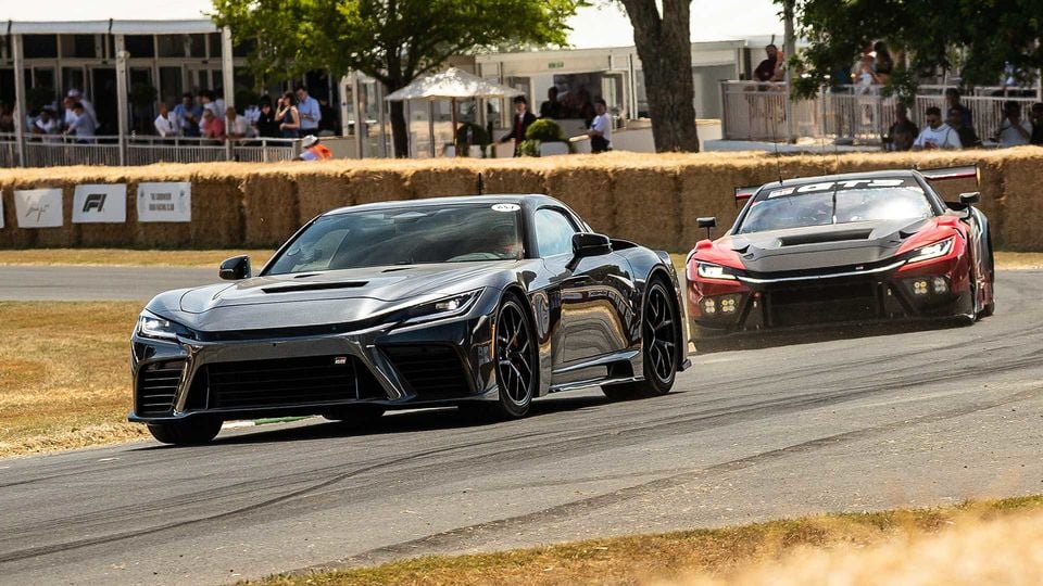A black Toyota GR GT supercar leads a red Toyota GR GT3 race car along the circuit at the Goodwood Festival of Speed, with hay bales and spectators visible in the background.