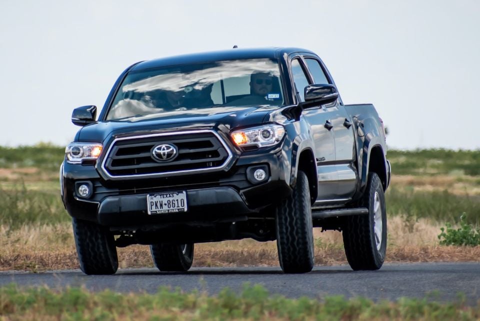 A dark Toyota Tacoma pickup truck seen from the front three-quarter angle, driving on a road through open grassland