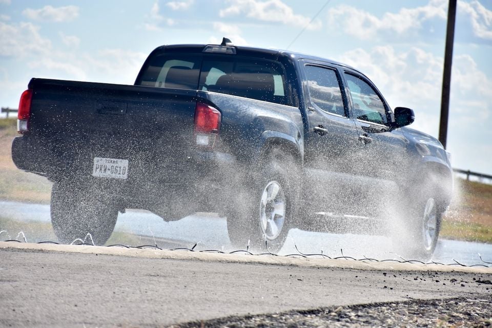 A dark-coloured Toyota Tacoma pickup truck seen from the rear-side, driving through a flooded road and throwing up a large spray of water