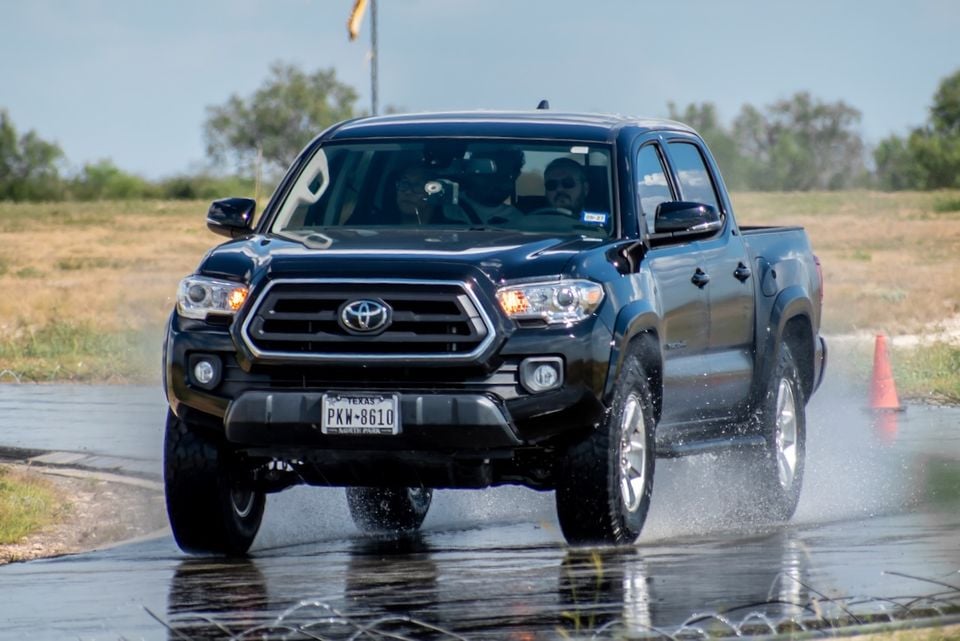 A dark Toyota Tacoma dual-cab ute driving through a wet handling course with orange cones visible, seen from the front three-quarter angle