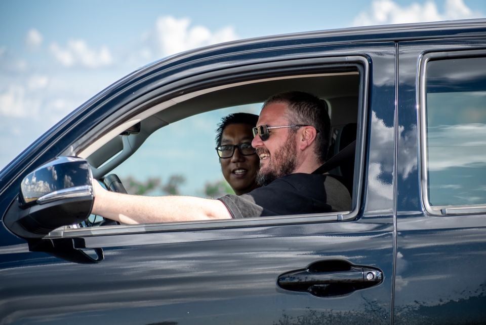 Two men sitting inside a dark blue ute, seen from the driver's side window, smiling while one holds the steering wheel