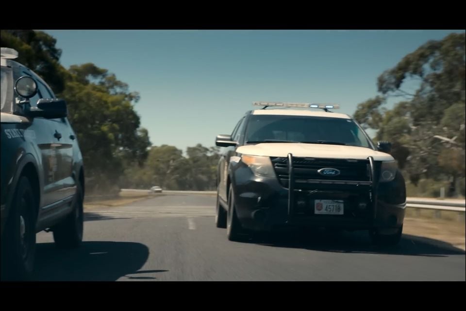 Two Ford police SUVs with roof-mounted light bars driving on a rural road lined with eucalyptus trees