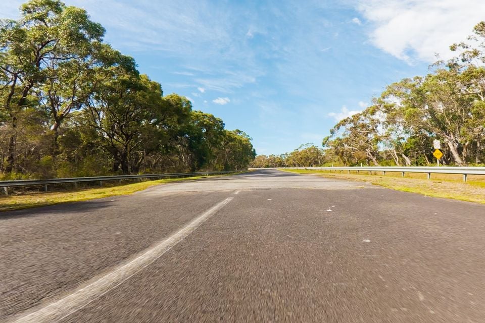 A low-angle view of an open two-lane Australian road lined with gum trees and guardrails under a blue sky