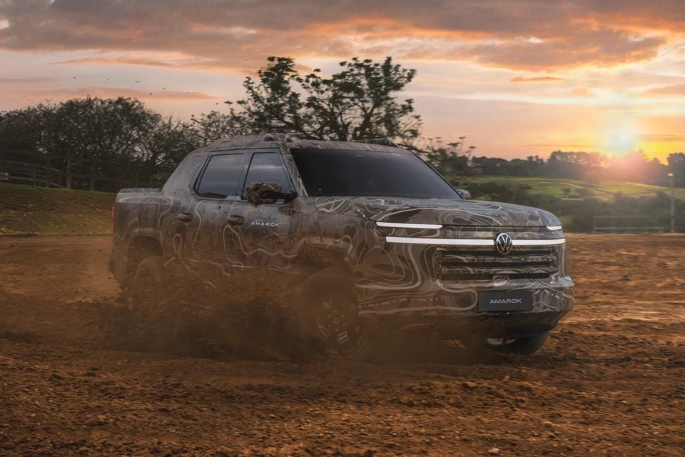A camouflaged Volkswagen Amarok ute driving through dirt, kicking up dust at sunset