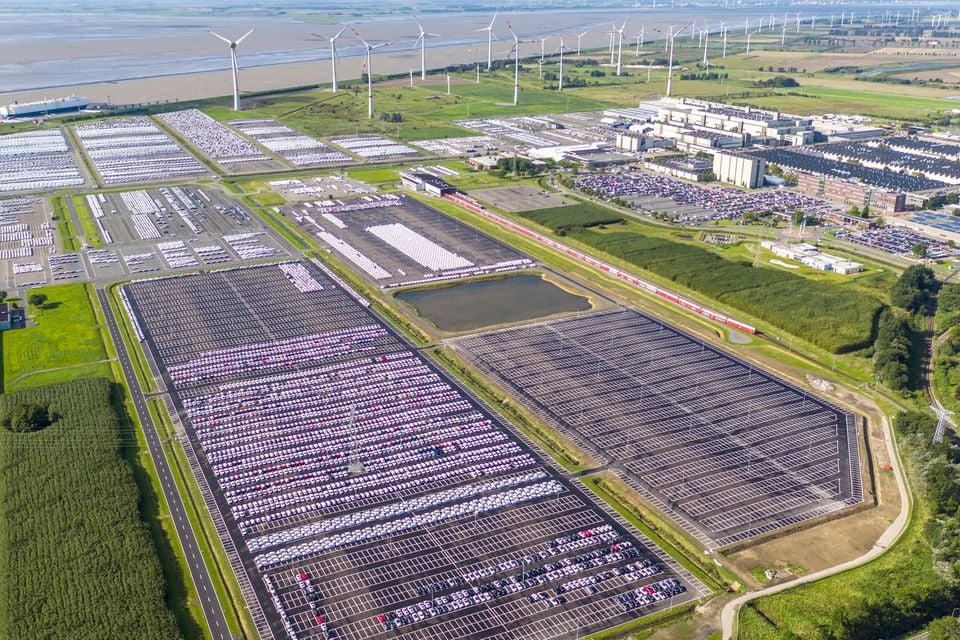 Aerial view of the Volkswagen Emden factory and vehicle storage lots, with thousands of new cars parked in rows across a large compound, wind turbines and flat green landscape visible in the background.