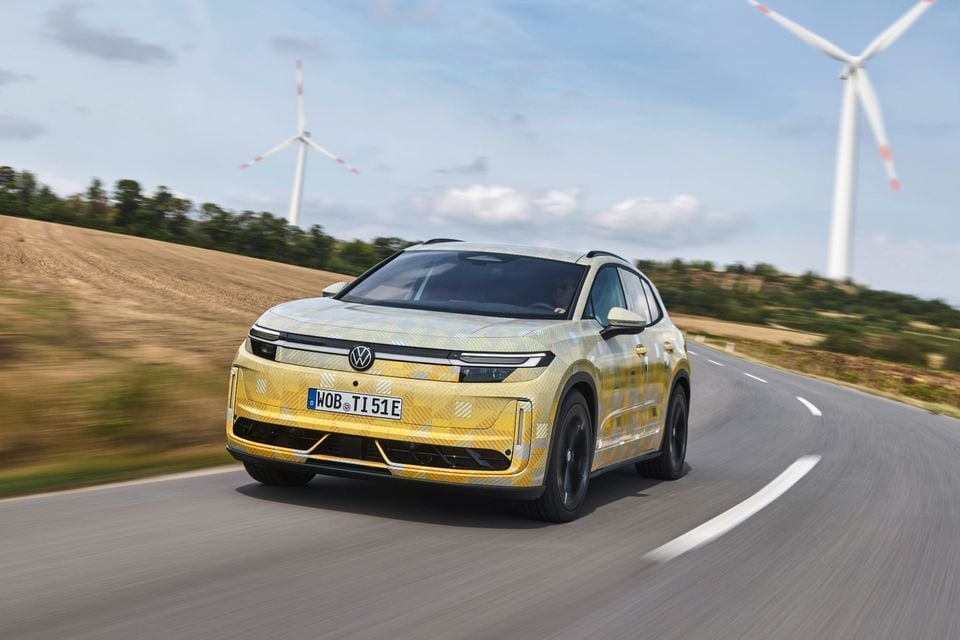 A camouflage-wrapped Volkswagen ID. Tiguan prototype SUV drives on a rural road past wind turbines.