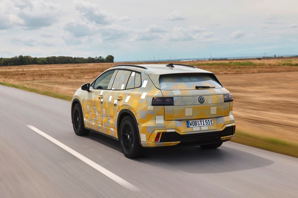 A camouflage-wrapped 2027 Volkswagen ID. Tiguan prototype SUV driving on a rural road, seen from the rear quarter.