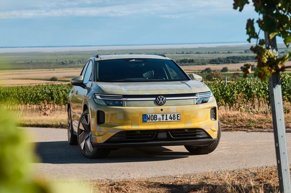 A camouflage-wrapped 2027 Volkswagen ID. Tiguan prototype SUV parked on a road beside a vineyard, viewed from the front.