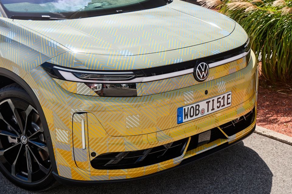 A Volkswagen ID. Tiguan prototype wrapped in yellow and grey camouflage, seen close up from the front showing its headlight and grille badge.