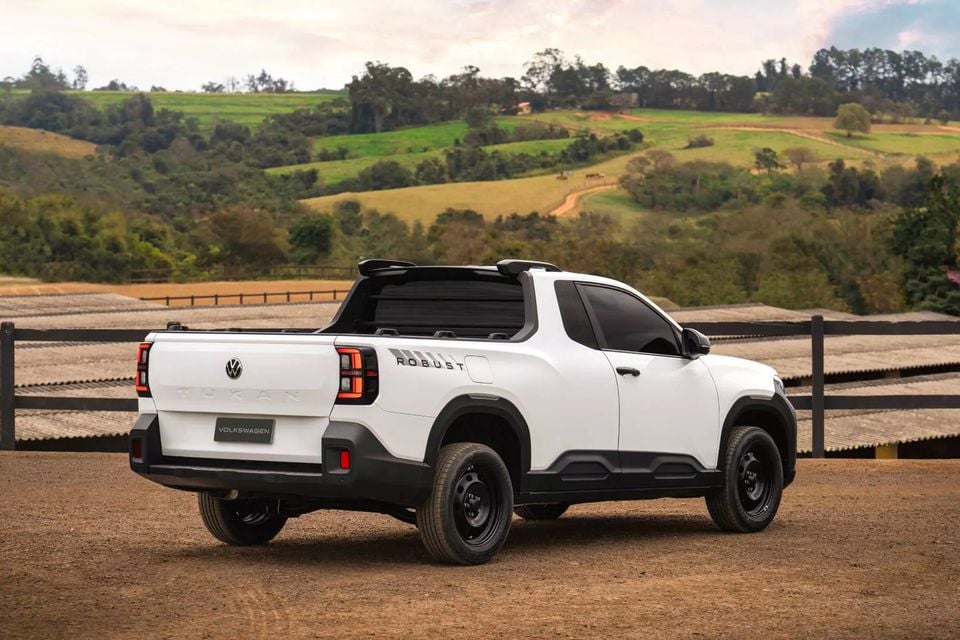 A white Volkswagen Tukan Robust ute seen from the rear three-quarter angle, parked on a dirt surface with rolling green hills in the background.