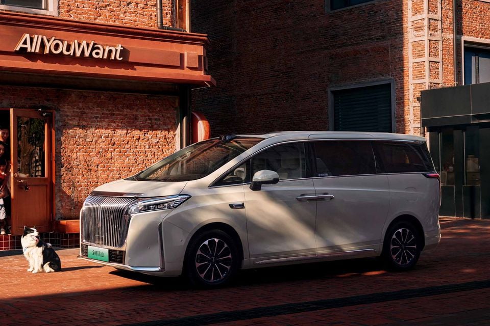 A cream-coloured WEY Gaoshan large minivan parked outside a brick building, seen from the front-side angle with a dog sitting nearby