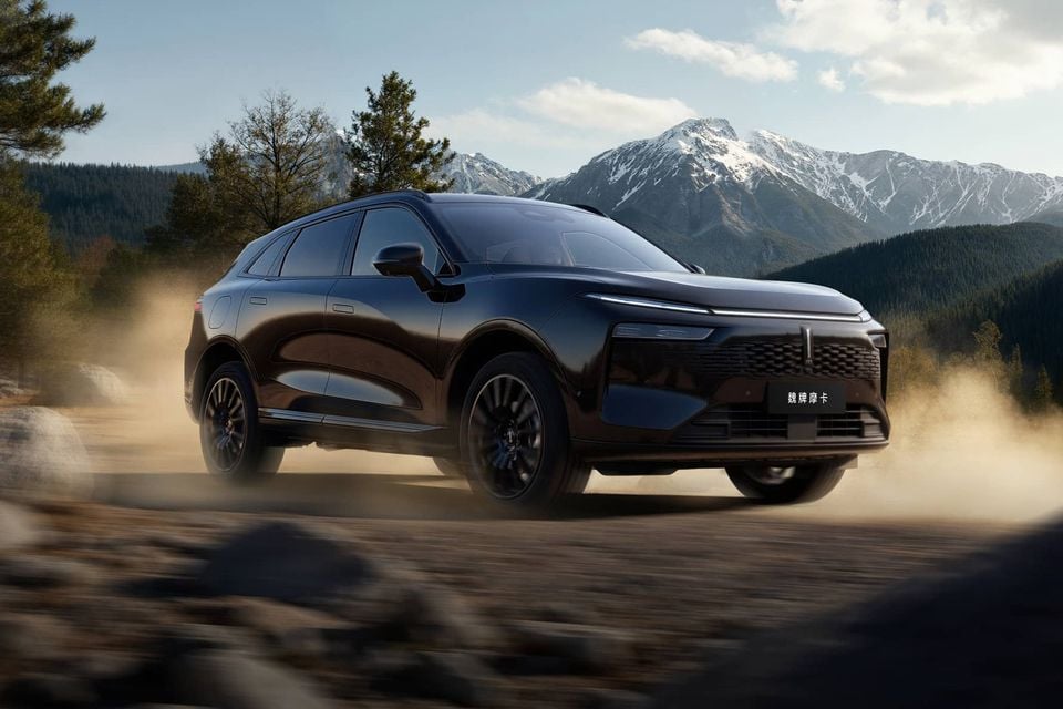 A black WEY Mocha SUV driving on a dusty gravel road with snow-capped mountains in the background, seen from the front-side