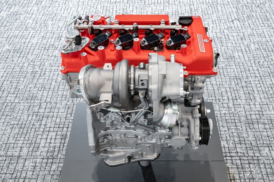 Um motor turboalimentado de quatro cilindros com uma tampa vermelha do came exibida em um suporte, mostrando o turboalimentador e acessórios