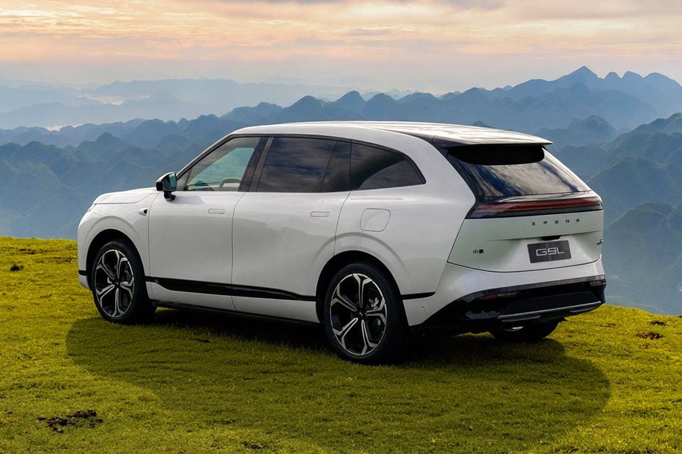 A white Xpeng G9L SUV seen from the rear three-quarter angle, parked on a grassy hillside with a mountainous landscape in the background.