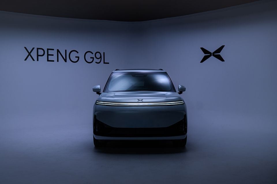 XPeng G9L SUV in grey seen from the front in a dimly lit studio, with its full-width LED light bar and XPeng branding on the wall behind