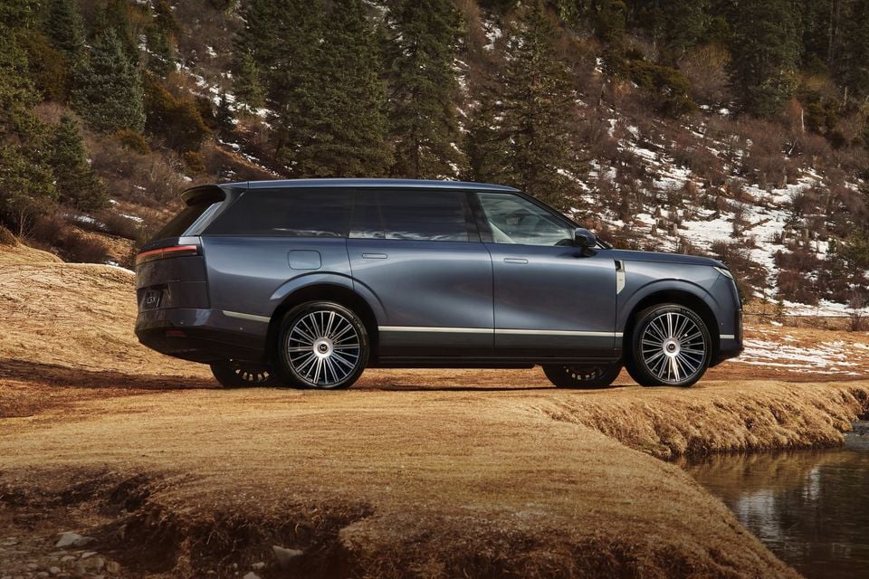 A grey Xpeng GX large SUV seen from the side, parked on a dirt track with a snow-dusted forested hillside in the background.