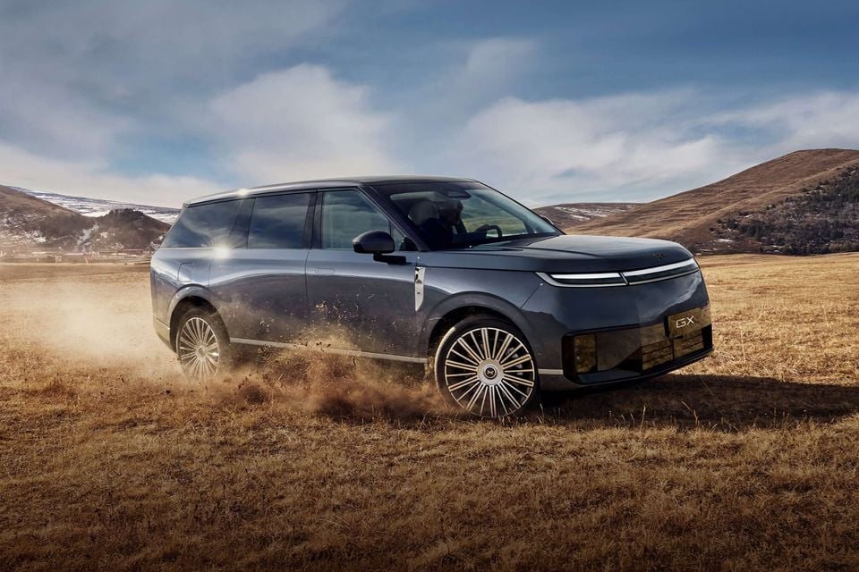 An Xpeng GX large SUV in dark blue driving through an open grassy field kicking up dust, seen from the front-side