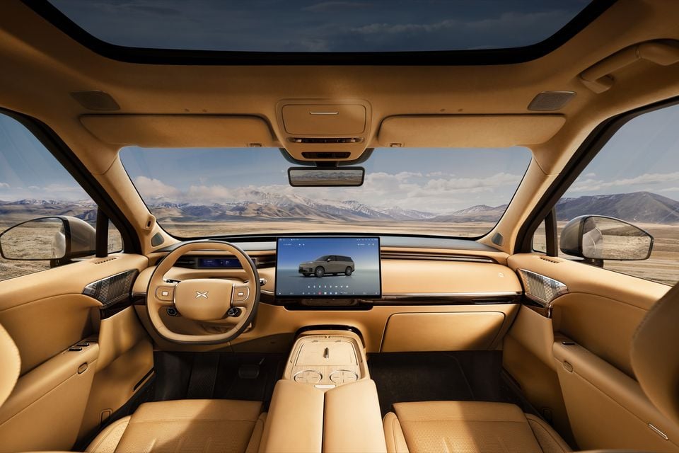 Interior of an Xpeng GX showing tan leather seats, a large central touchscreen infotainment display, and a flat-bottom steering wheel with an X logo, set against a desert mountain backdrop.