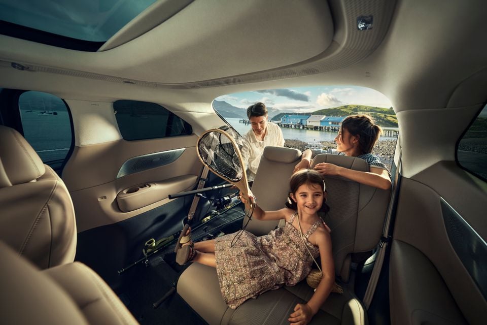 Interior of an XPENG X9 SUV showing the spacious rear cabin with cream leather seats, a panoramic roof, and a young girl holding a fishing net while two adults load fishing gear through the open tailgate, with a coastal scene visible in the background.