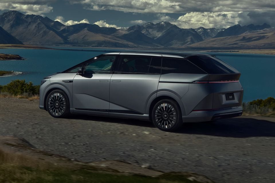 A silver Xpeng X9 large MPV seen from the rear three-quarter angle, driving along a scenic road beside a mountain lake