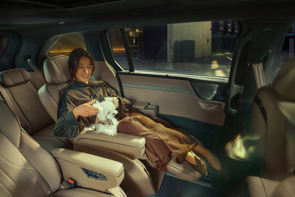 A woman relaxes in the luxurious rear seat of an Xpeng X9, smiling as she holds a small white dog on her lap, surrounded by premium beige leather upholstery and extended footrest.
