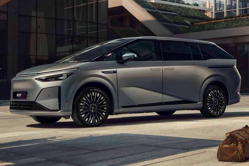 A silver Xpeng X9 electric minivan seen from the front-side angle, parked outside a modern building