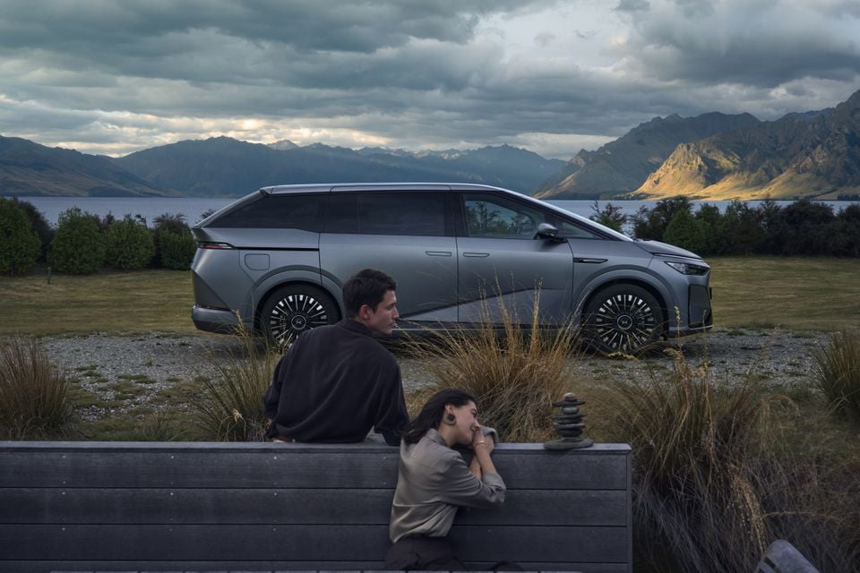 A grey XPENG X9 large electric MPV parked by a scenic lake and mountain backdrop, with two people sitting on a bench in the foreground