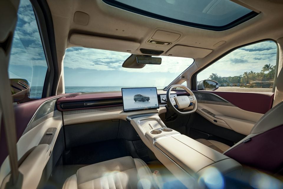 Interior of the Xpeng X9 showing the front cabin with a large central touchscreen, yoke-style steering wheel, cream and burgundy upholstery, and a panoramic sunroof, parked near a beach