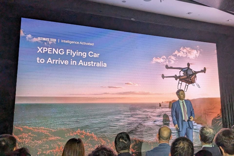 A presenter at an XPENG event stands in front of a large screen announcing 'XPENG Flying Car to Arrive in Australia', with an audience visible in the foreground.