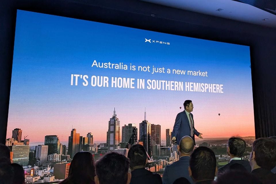 An Xpeng presenter speaks on stage at an Australian launch event, with a large screen behind him displaying the message 'Australia is not just a new market – IT'S OUR HOME IN SOUTHERN HEMISPHERE' over a Melbourne city skyline.