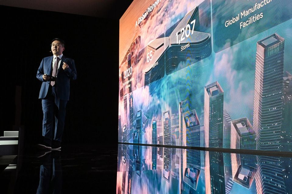 A presenter speaks on stage at an XPeng event in Melbourne, with a large screen displaying global brand statistics including 1,207 stores and manufacturing facilities.