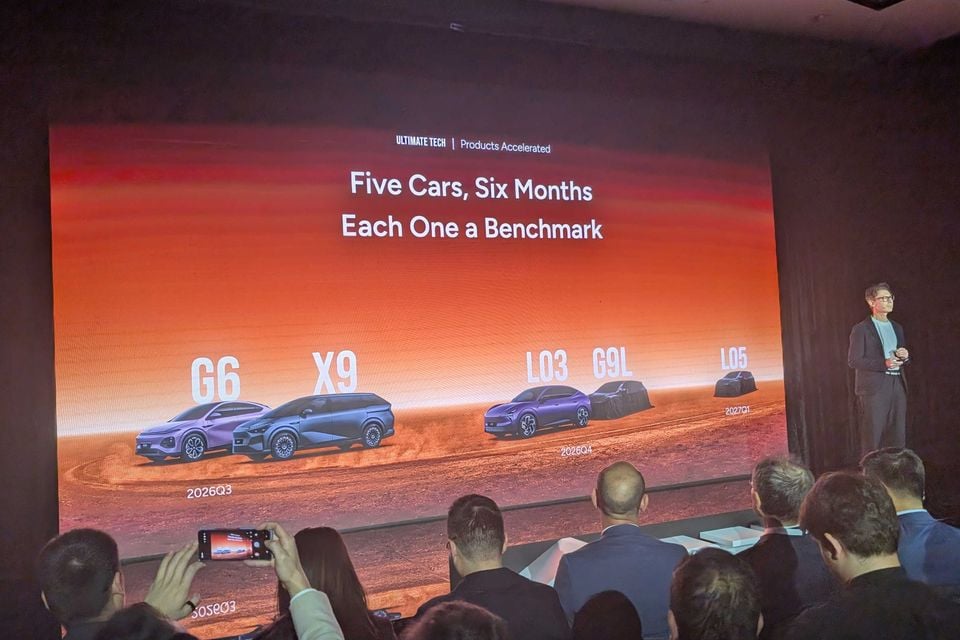 A presenter speaks at an XPENG product event, with a large screen showing five upcoming models — G6, X9, L03, G9L, and L05 — under the headline 'Five Cars, Six Months, Each One a Benchmark'