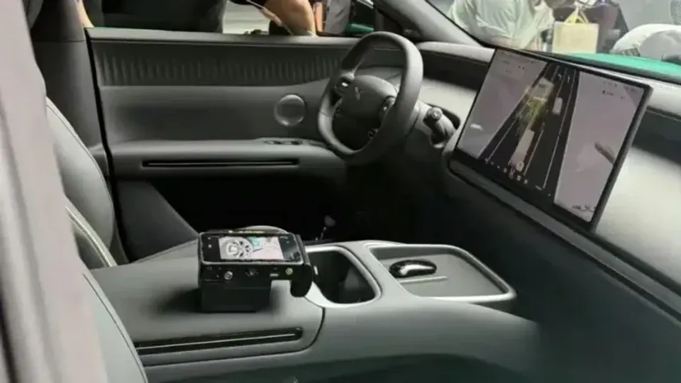 Interior of the Xpeng Mona L05 showing the dashboard, yoke-style steering wheel, large infotainment screen displaying navigation, and a small secondary control device resting on the centre console