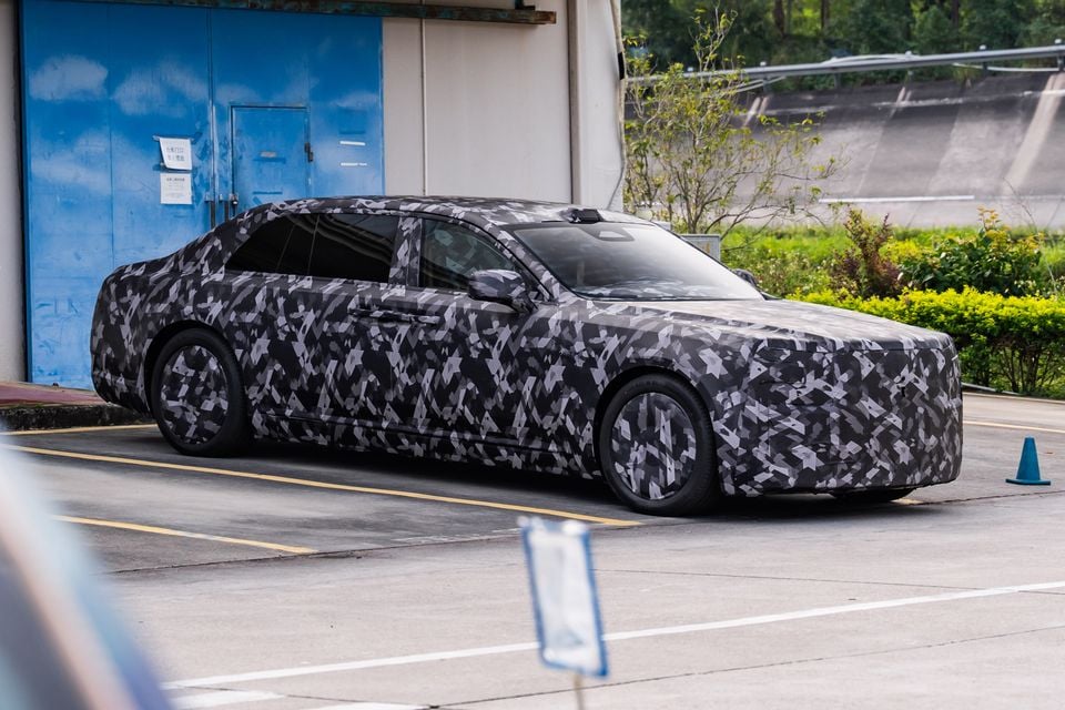 A large sedan wearing black and grey camouflage wrap is seen testing outside a facility, viewed from the front and side.