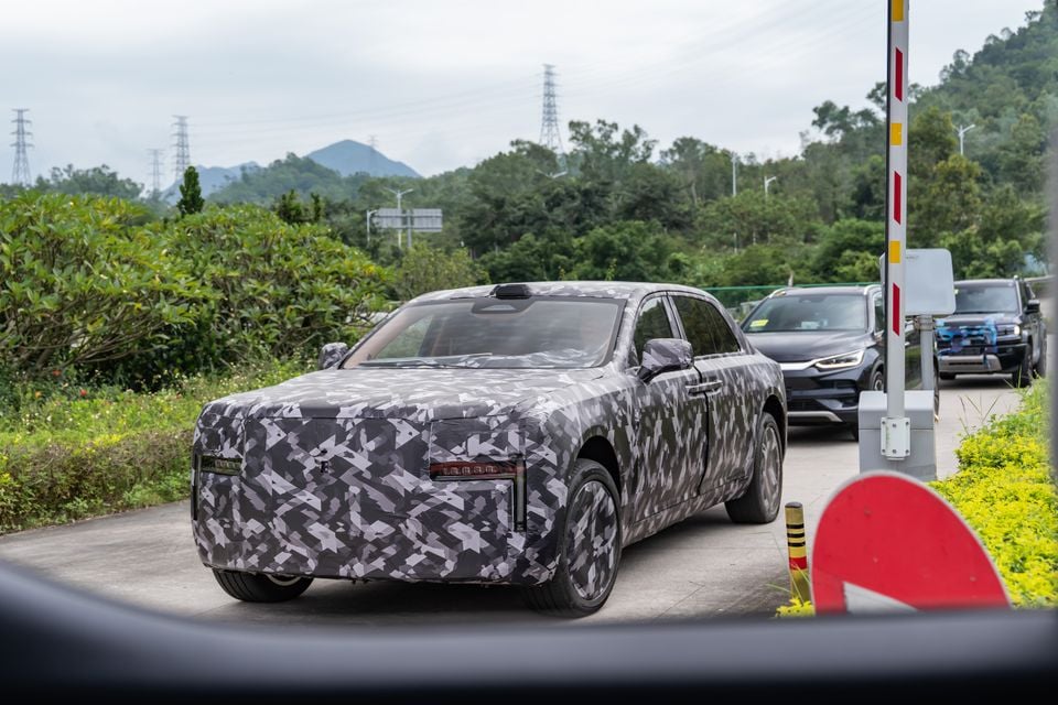 A camouflage-wrapped prototype sedan, likely a Rolls-Royce, seen from the front quarter on a test track with other vehicles behind it.
